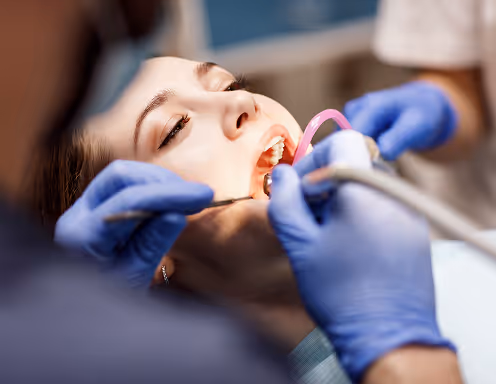 Dental professional wearing blue gloves performing oral cleaning on a patient's open mouth using dental tools.