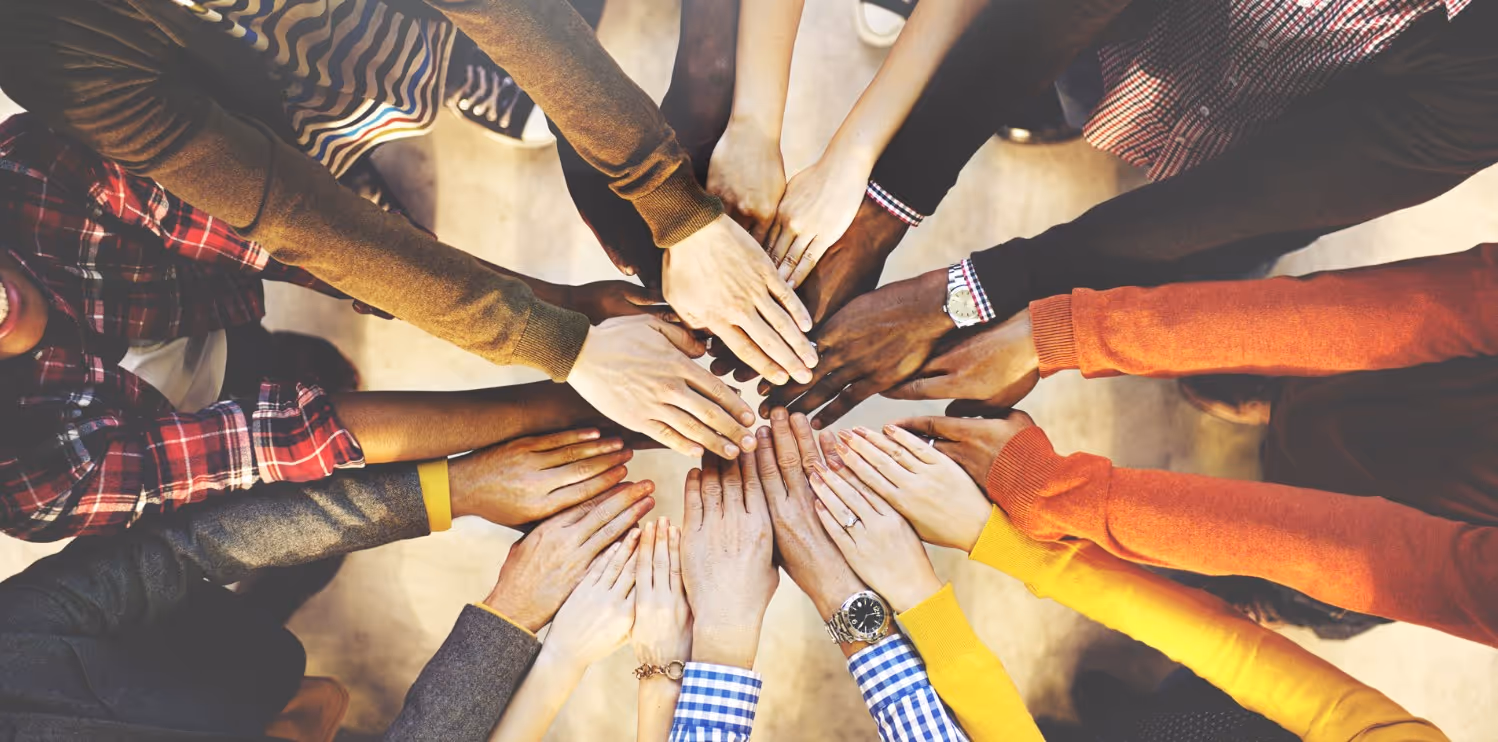 Multiple people of diverse skin tones and attire placing their hands together in the center, symbolizing teamwork and unity.