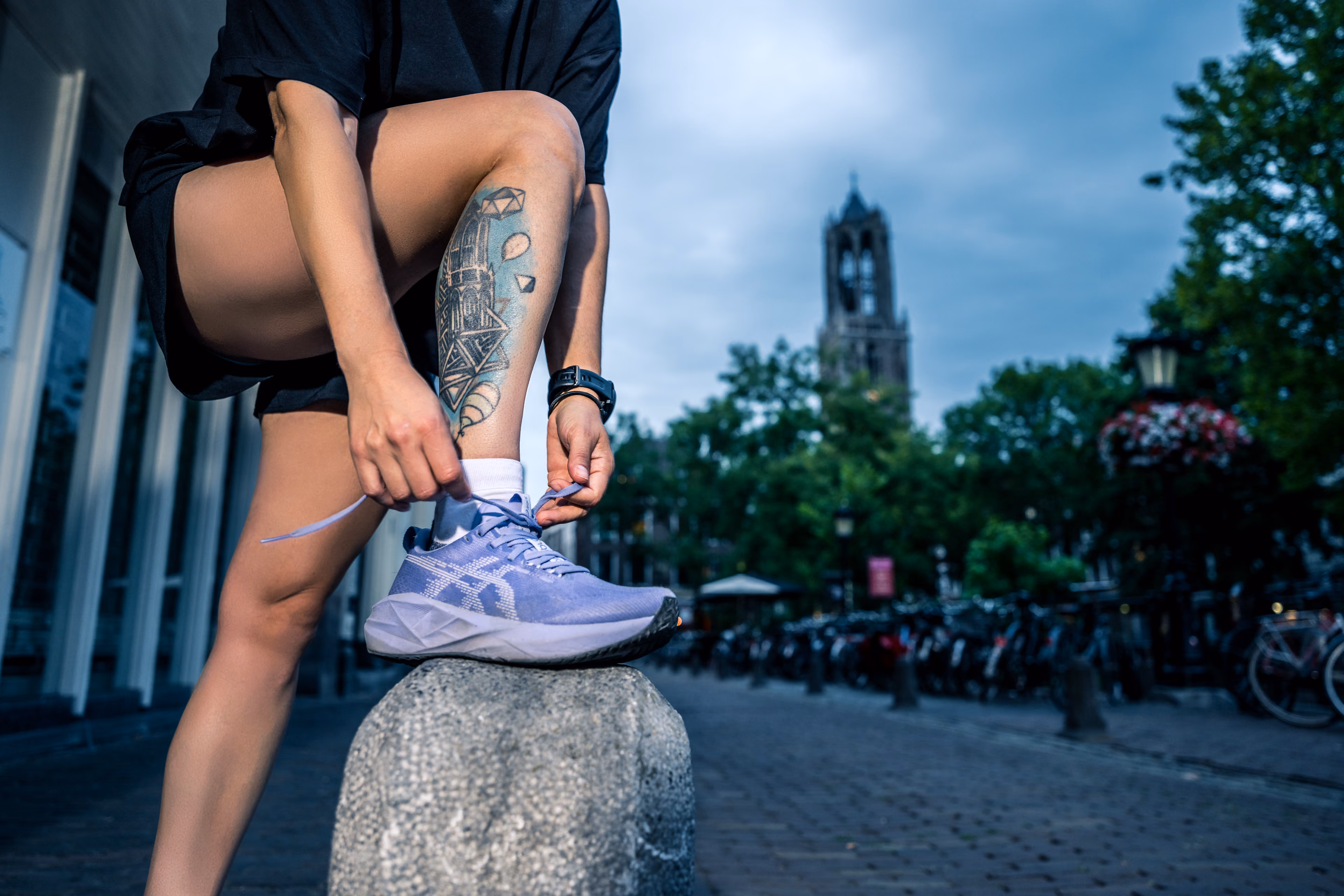A person wearing a pair of purple sneakers with a city in the background during the evening, with a detailed tattoo visible on the tibia.