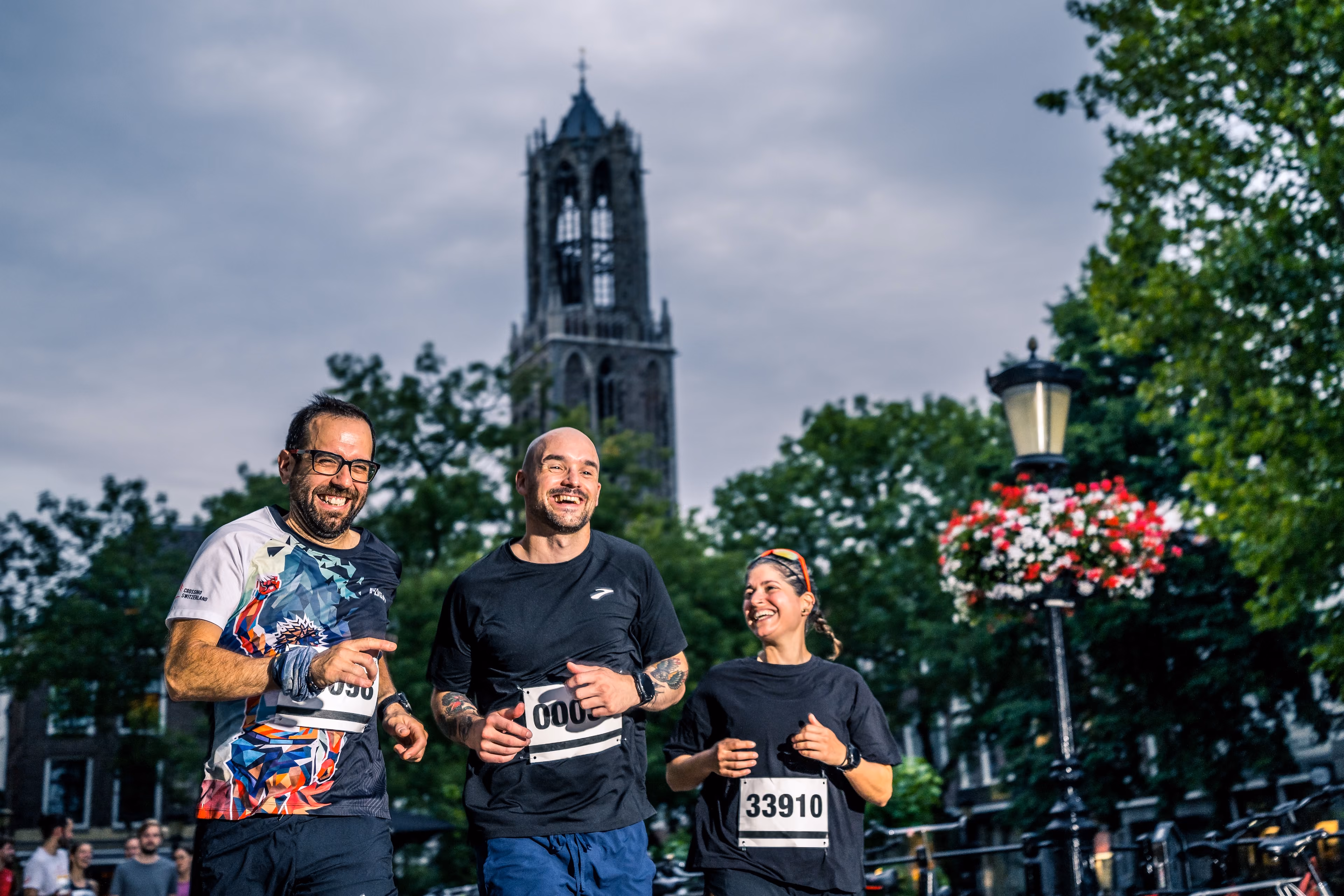 Three runners with race numbers laughing as they run through a city park with trees and a tall Gothic tower in the background under a cloudy sky.