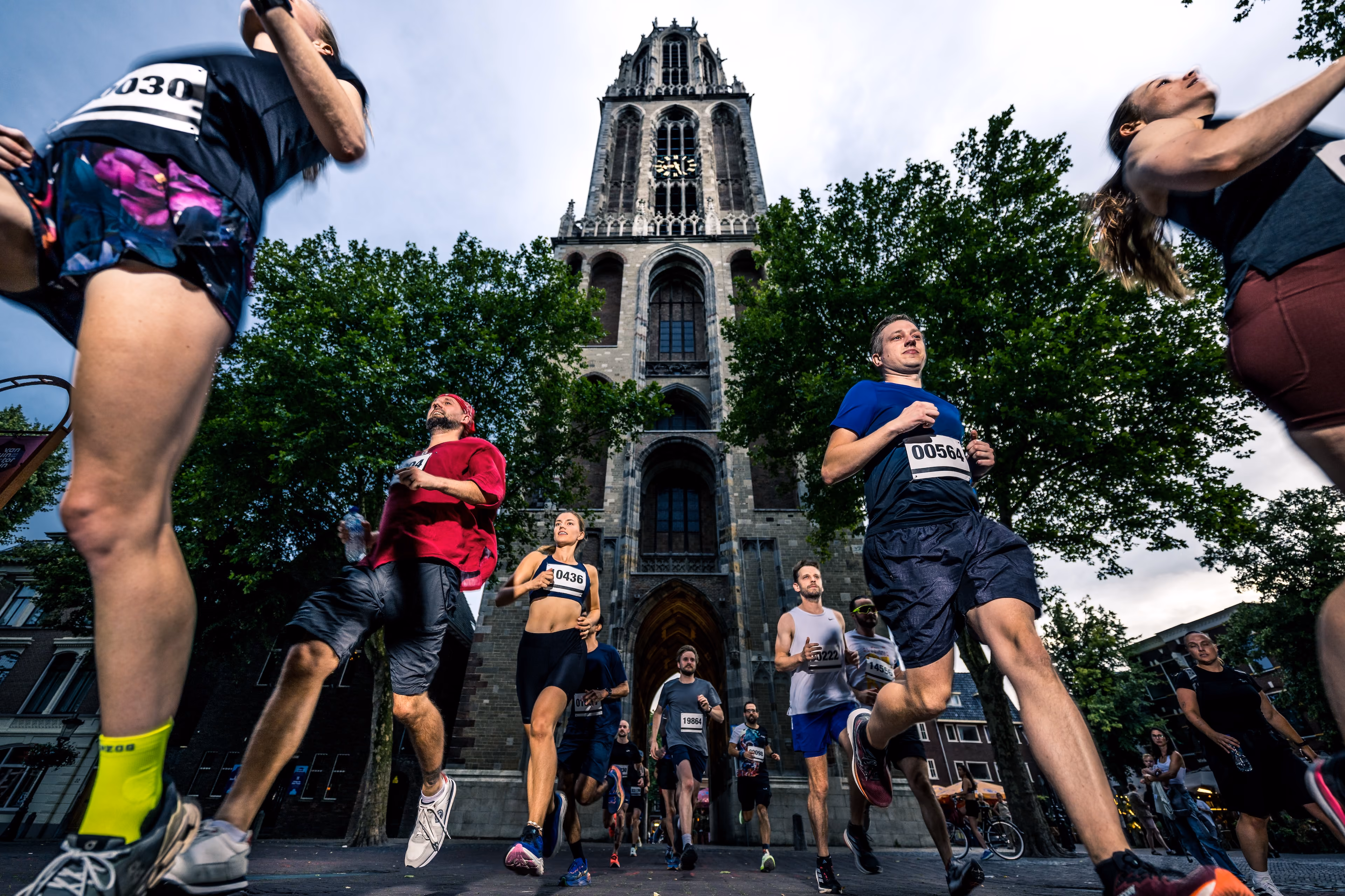Groep hardlopers loopt een straat met bomen in, met op de achtergrond een grote gotische kerktoren onder een bewolkte hemel.