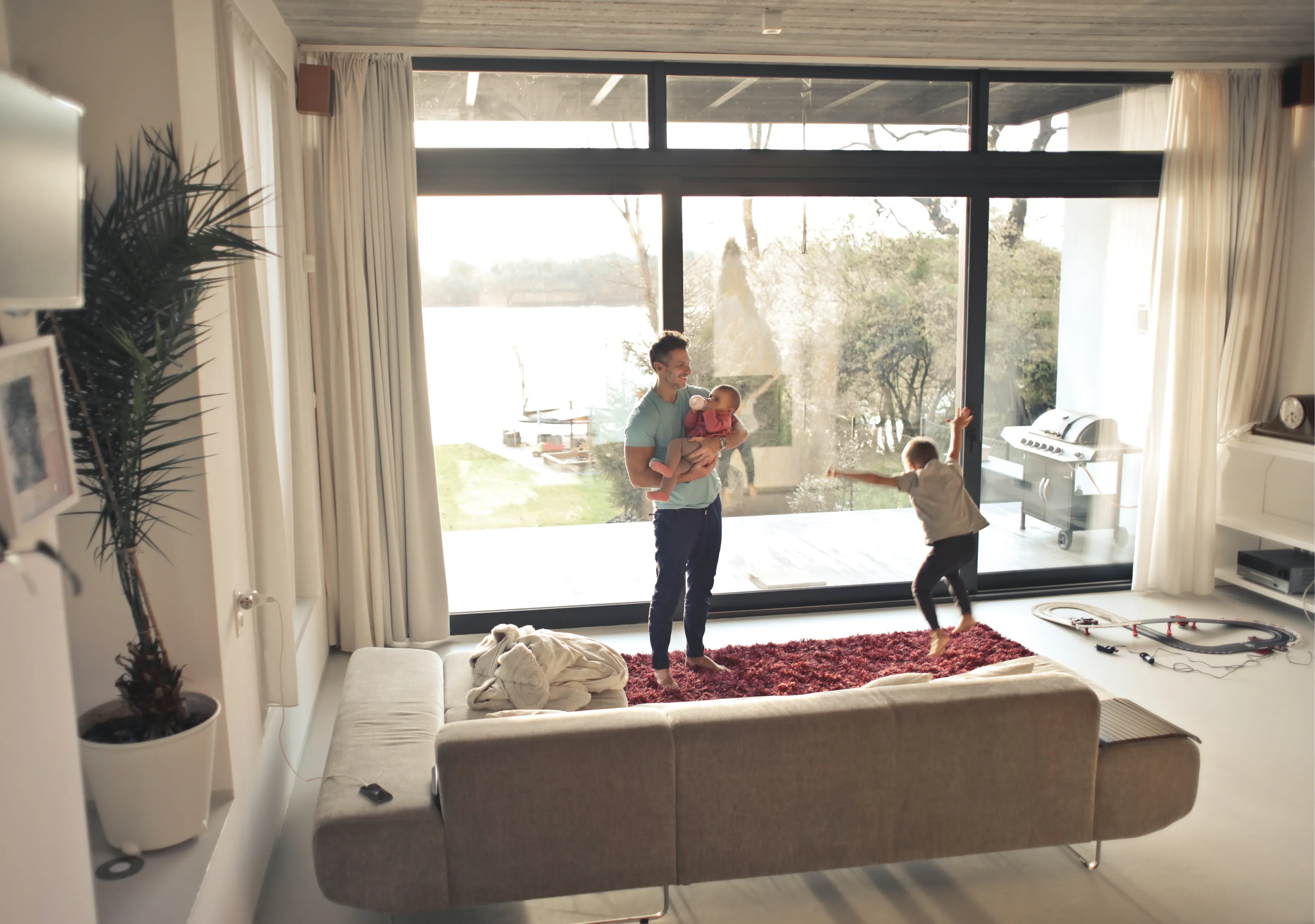 Father holding a baby and playing with a young child jumping on a red rug in a living room with large floor-to-ceiling windows overlooking a garden.