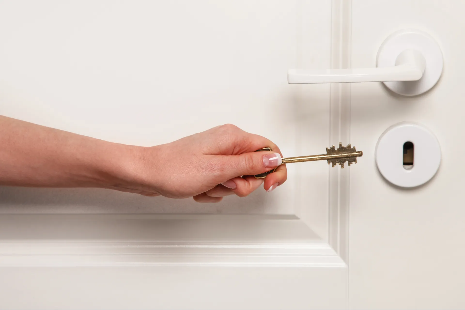 Hand holding a brass key near the keyhole of a white door with a white handle.