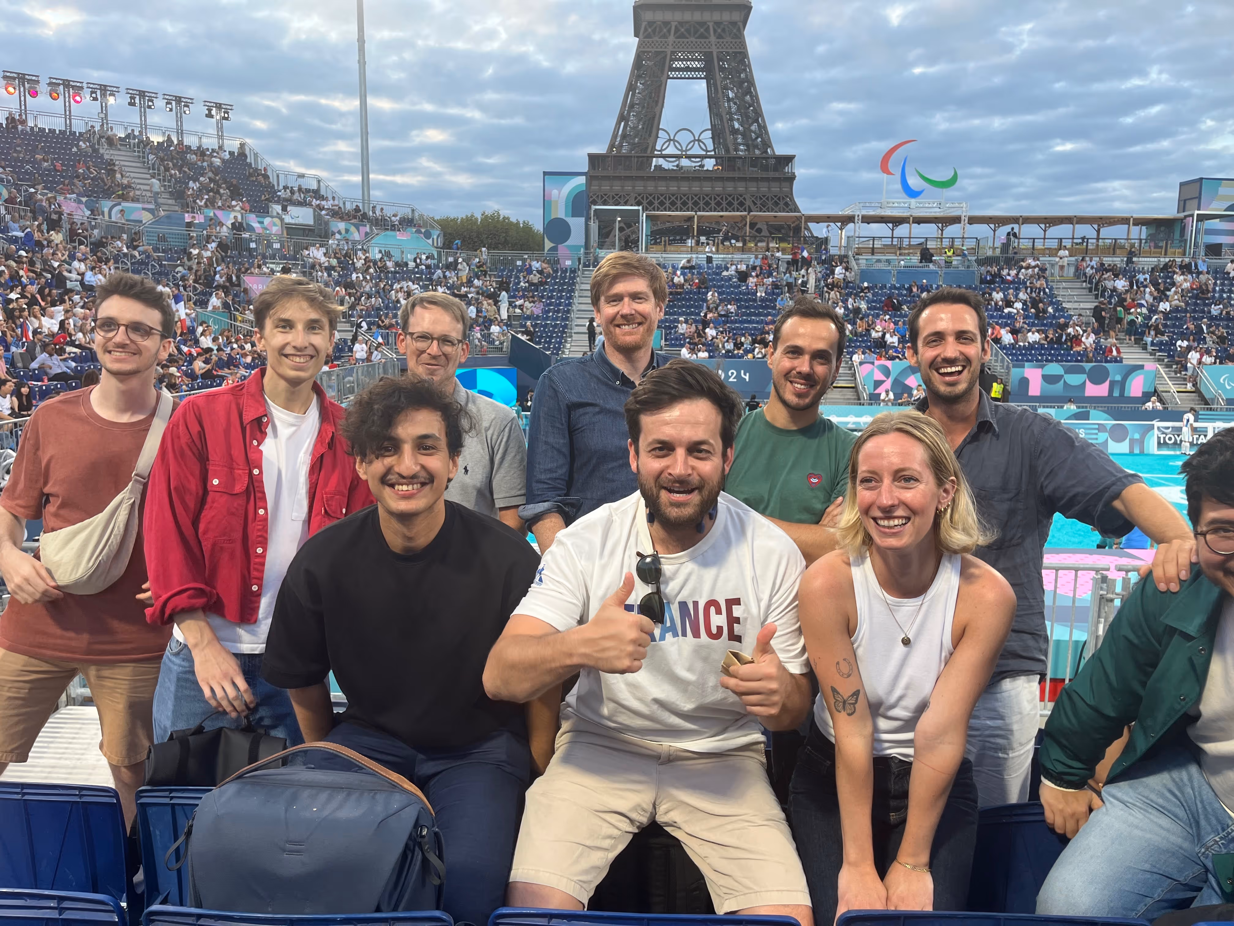 Group of ten smiling young adults posing in front of the Eiffel Tower and a stadium crowd during a sporting event.