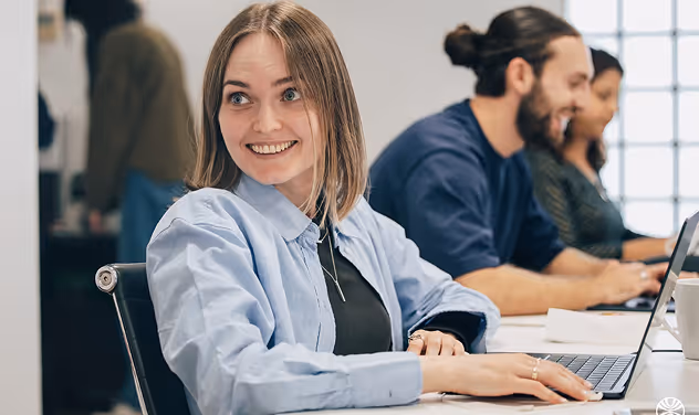 Smiling woman with laptop, two colleagues work beside her in a bright office.