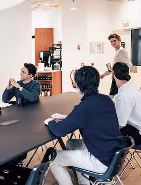 Four men in a modern office, three seated at a table, one walking with a cup in hand.