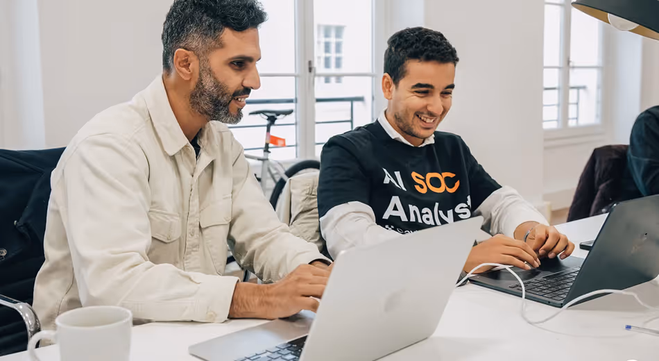 Two men sitting at a desk, working on laptops and smiling in a bright office.