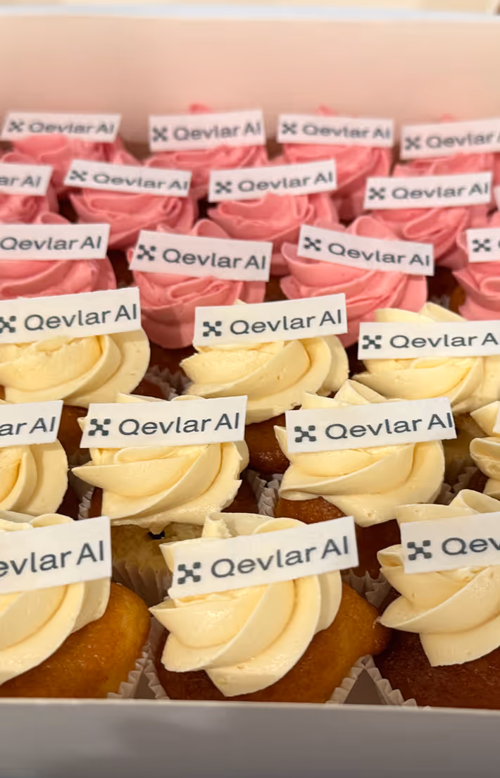Box of cupcakes with pink and cream swirled frosting topped with Qevlar AI logo flags.