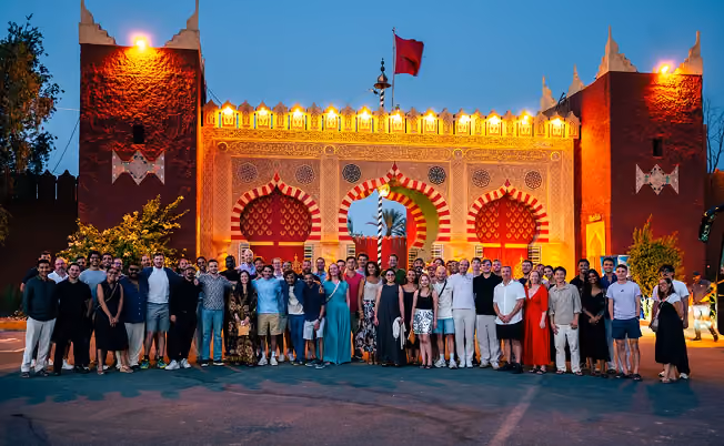 A large group of people standing together posing for a photo in front of a traditional Moroccan-style gate illuminated with warm yellow lights at dusk, with a red flag on top and two tall red towers on each side.
