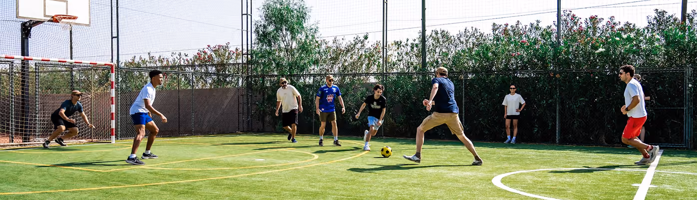 Seven men playing soccer on a small outdoor field with artificial turf, surrounded by a fence and greenery, with one player about to kick a yellow and black ball near the goal guarded by a crouching goalkeeper.