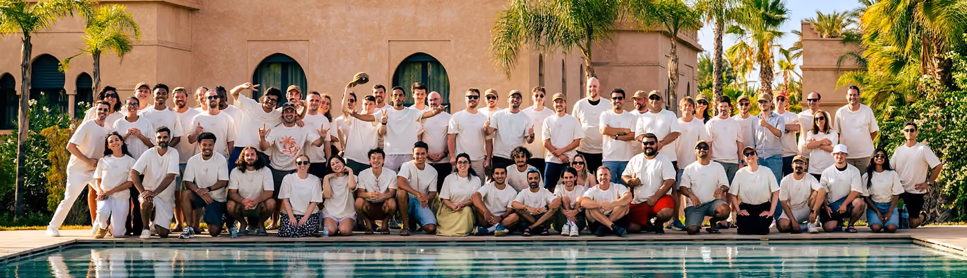 Large group of people wearing matching light-colored shirts posing by a pool with palm trees and a beige building in the background.
