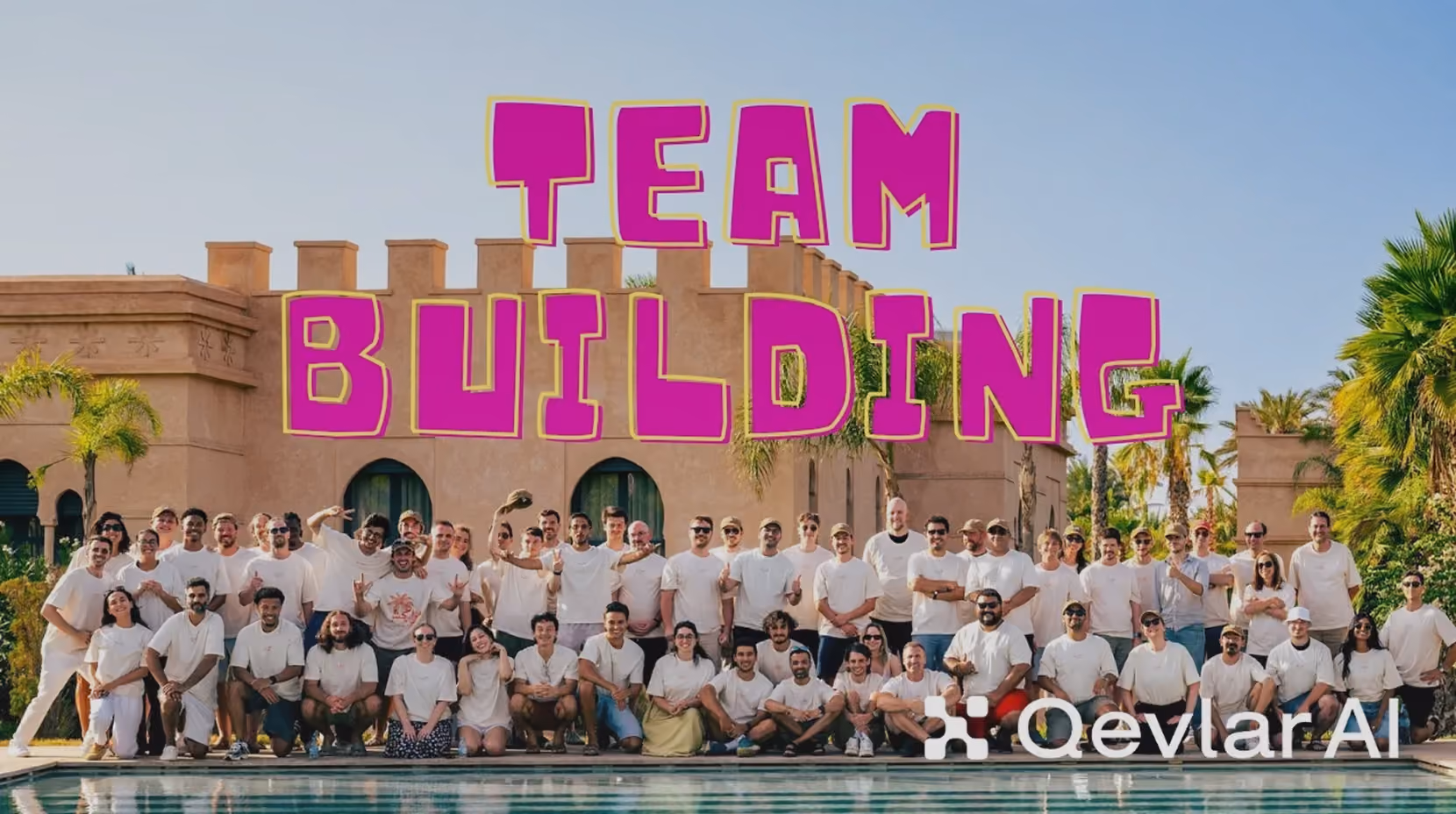 Large group of people posing by a pool in front of a tan building with palm trees, wearing mostly white shirts, with bold magenta text above reading 'TEAM BUILDING'.