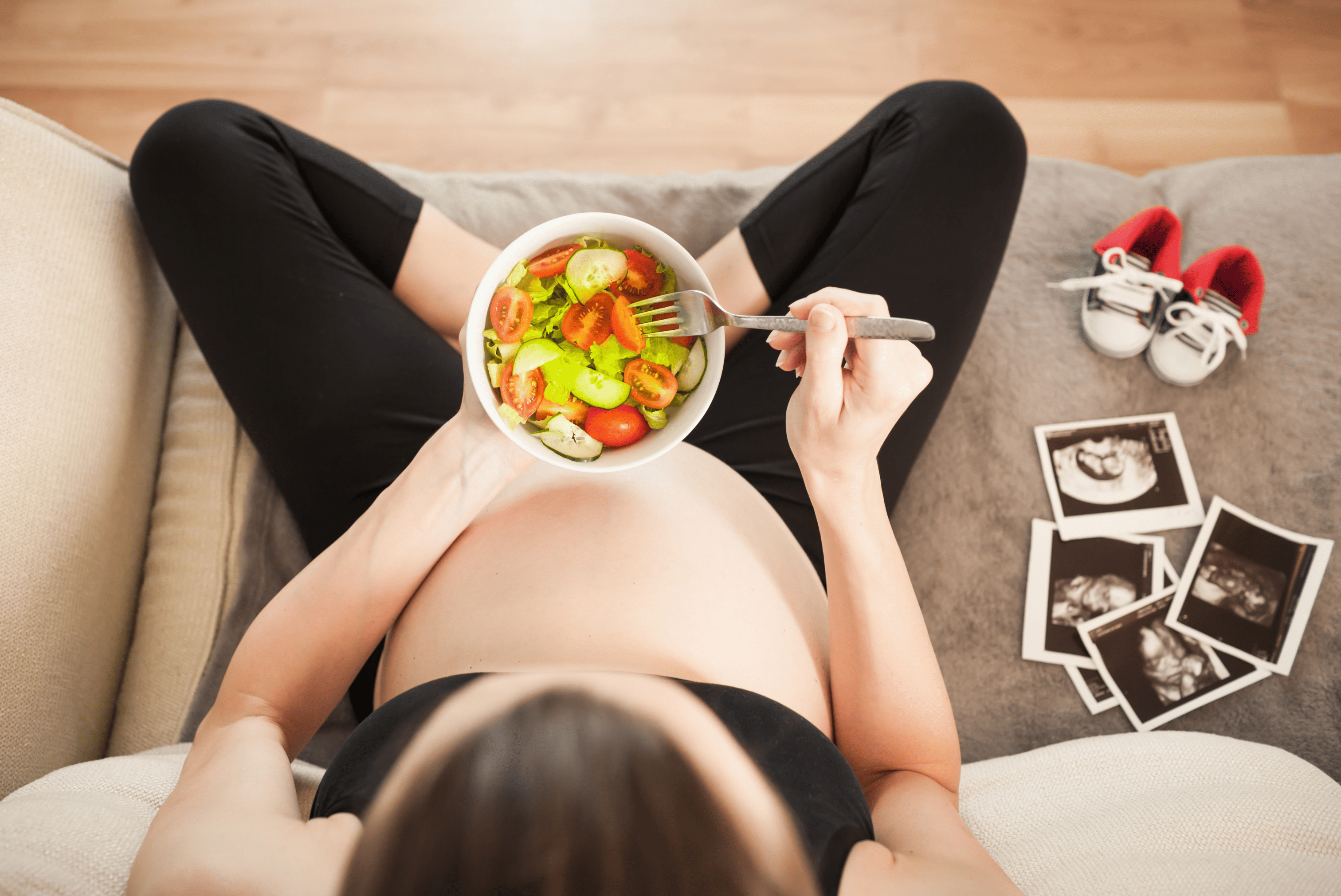 Femme enceinte assise mangeant une salade fraîche, avec échographies et chaussons de bébé à proximité, illustrant une alimentation saine pendant la grossesse.
