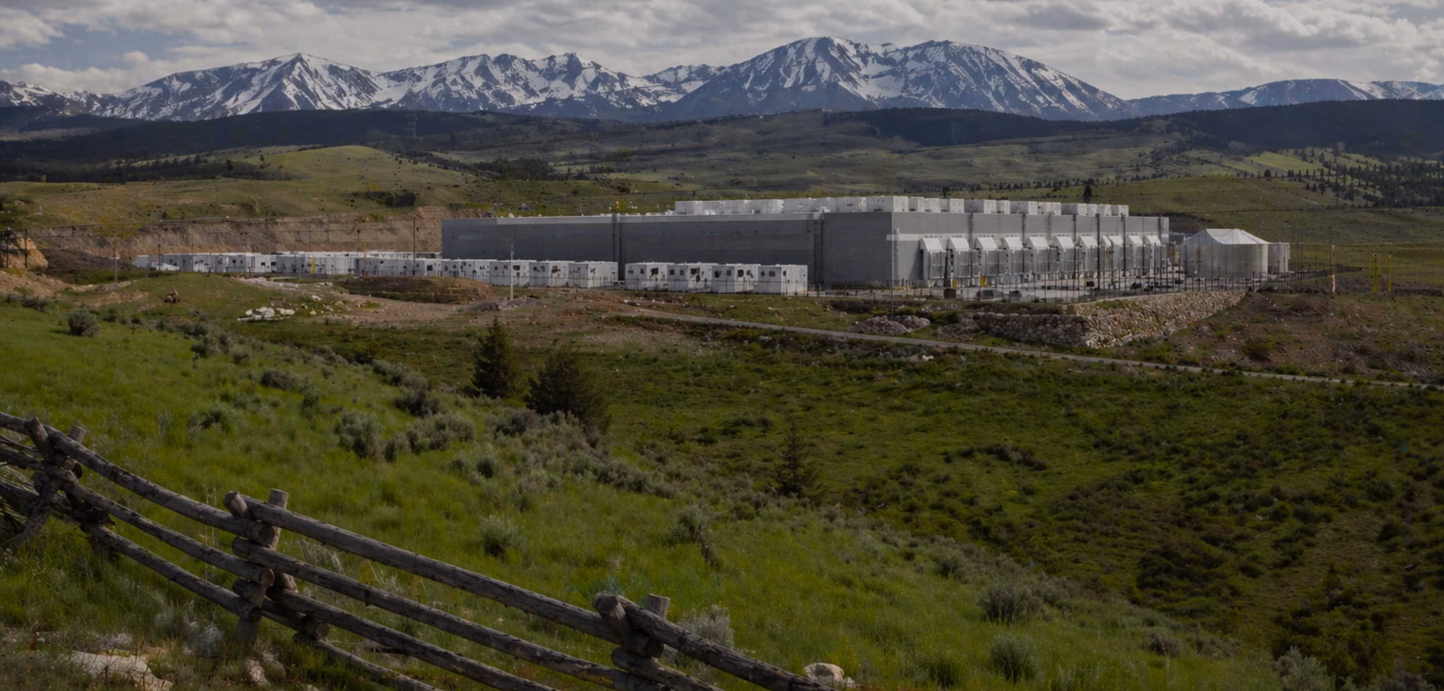 Large industrial building surrounded by green fields and mountains with snow patches in the background.