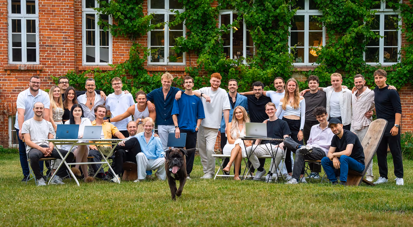 Gruppenfoto von 24 jungen Erwachsenen im Garten vor einem ziegelgedeckten Haus mit grünen Efeupflanzen, einige sitzen an Tischen mit Laptops, ein schwarzer Hund läuft im Vordergrund.