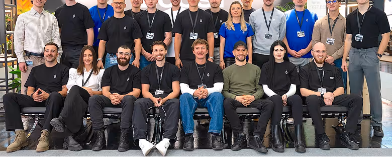 Gruppenfoto von 20 Personen, die in zwei Reihen vor einer modernen Innenraumkulisse posieren, viele tragen schwarze T-Shirts mit einem kleinen Logo.