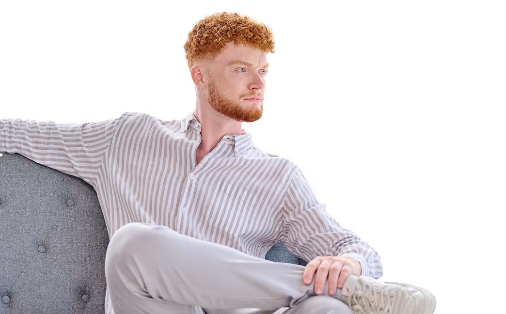 Junger Mann mit rotem lockigem Haar und Bart, lächelnd, trägt ein gestreiftes Hemd.
