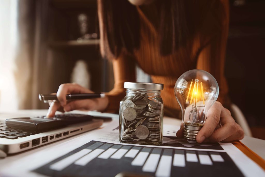 a woman using her calculator while holding a light bulb, and there is a jar of coins on her desk