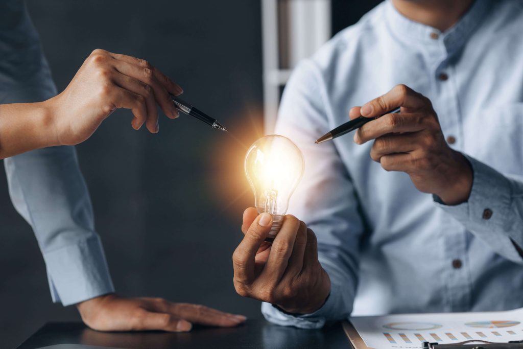 2 people looking at a light bulb and pointing at it with their pens