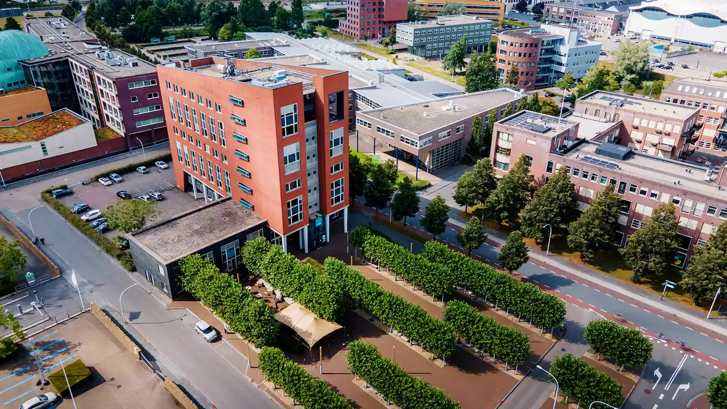 Luchtfoto van een modern kantoorgebouw met een roodbruine gevel, omringd door groen en parkeerplaatsen.
