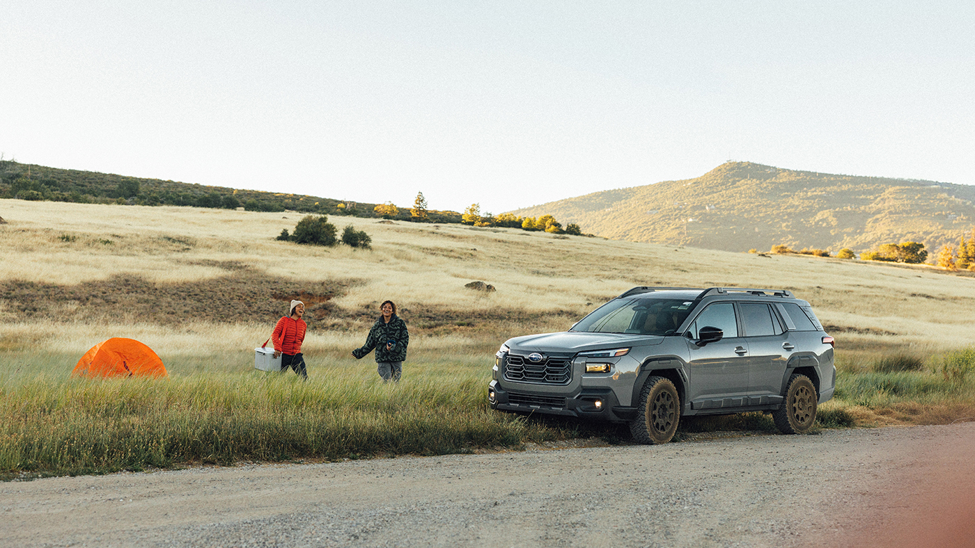 riginal unretouched automotive lifestyle photograph of a grey Subaru SUV parked next to a grassy field with two campers and an orange tent, showing the initial gravel road foreground and a blurred lens obstruction prior to professional retouching.