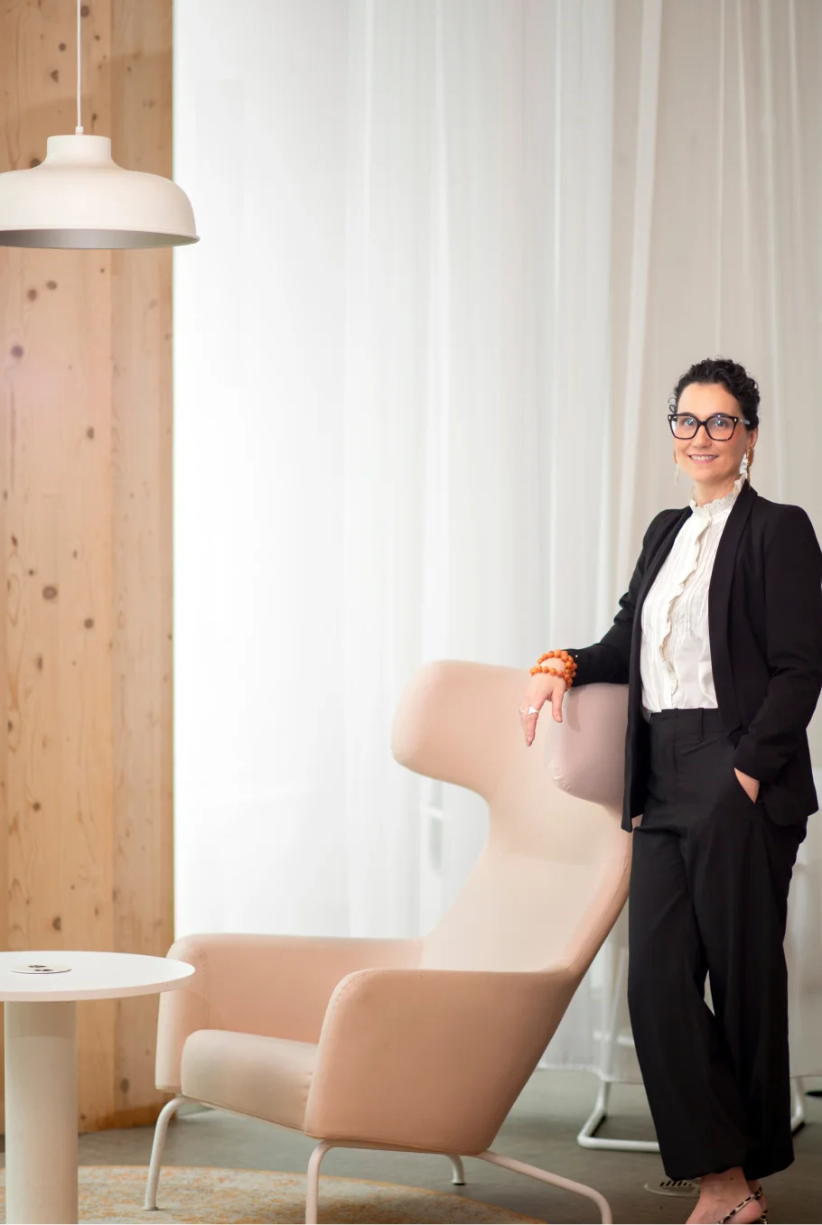 Mujer vestida con traje negro y blusa blanca, apoyada en un sillón rosa claro en un espacio con fondo de cortinas blancas y pared de madera.