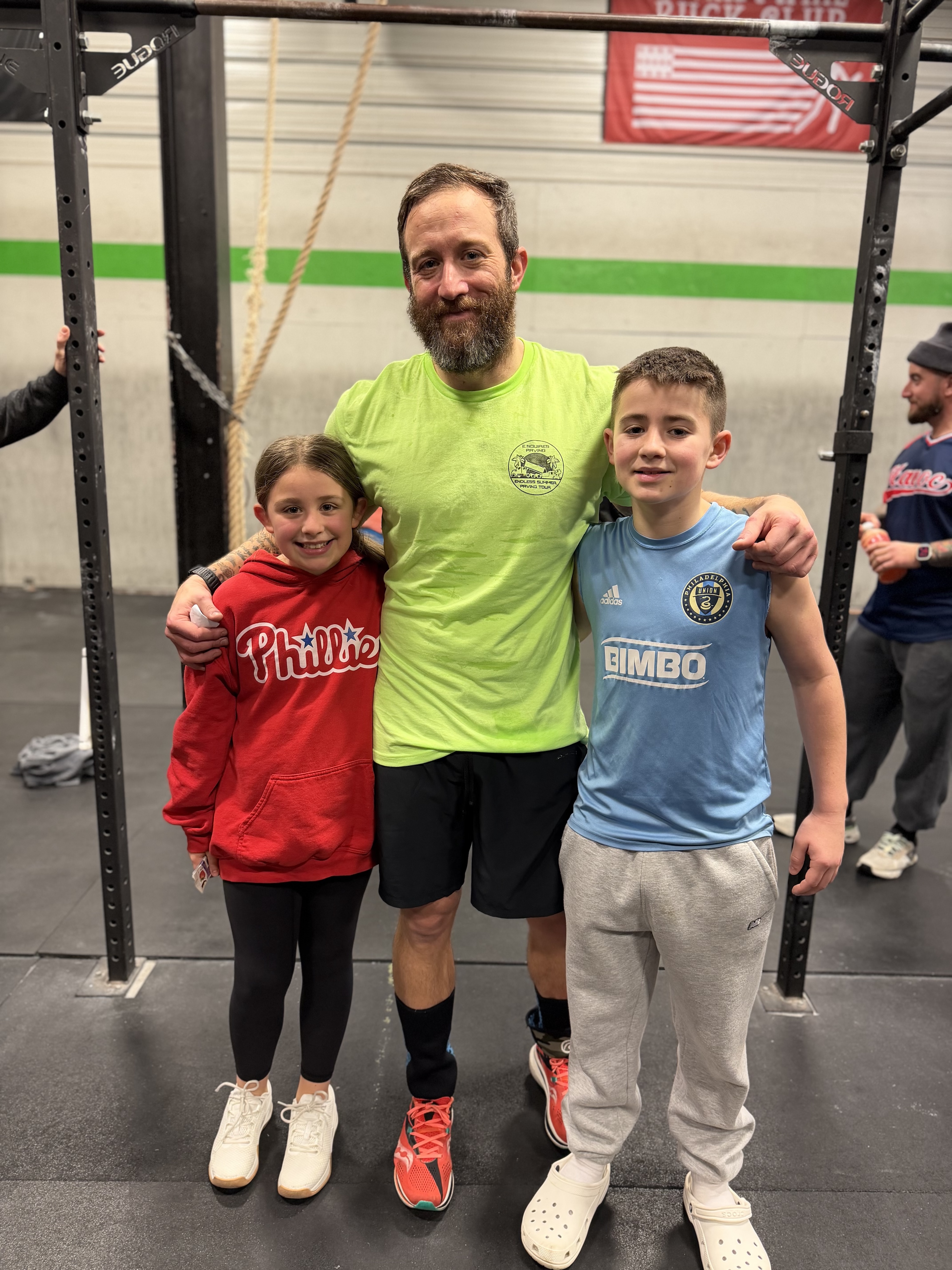 Man in a neon green shirt with arms around two children; girl in red Phillies hoodie and boy in blue Philadelphia Union soccer jersey, standing in a gym.