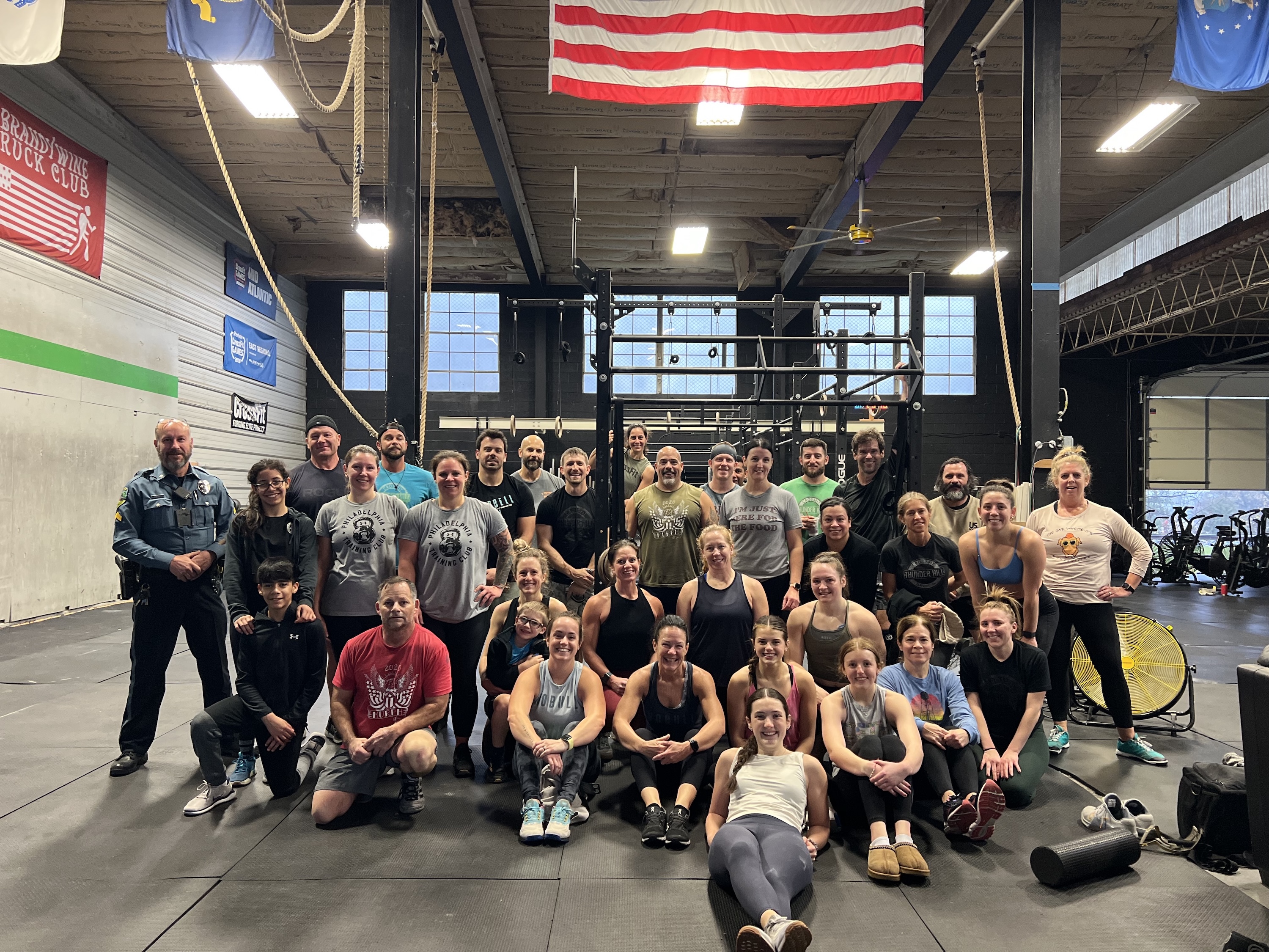 Group photo of around 30 diverse adults and children in workout attire posing inside a gym with flags and fitness equipment in the background.
