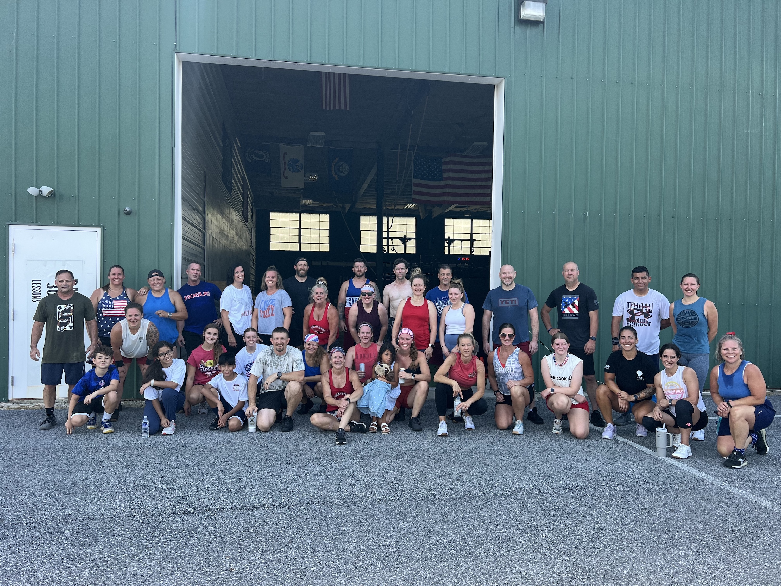 Group of diverse men, women, and children posing outside a green building with an open garage door.