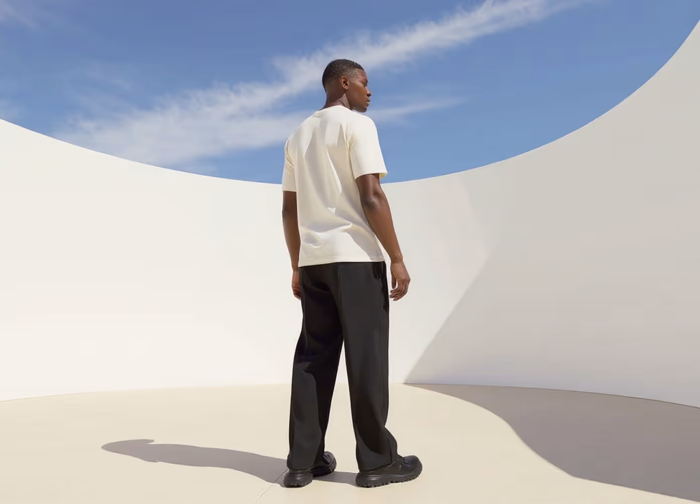 Man in cream t-shirt and dark trousers standing in a sunlit white courtyard, viewed from behind
