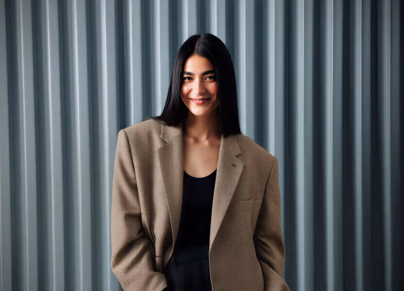 Smiling woman with long dark hair in a brown plaid blazer against a blue ribbed studio backdrop