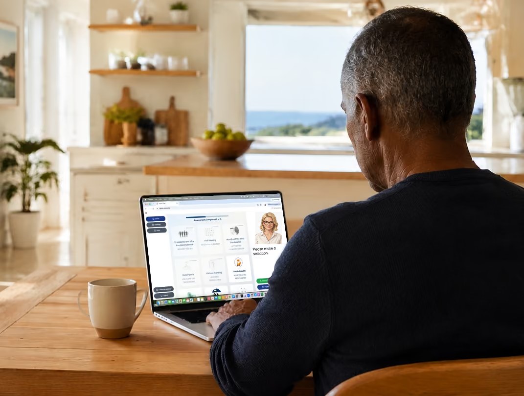 Older man at his kitchen table taking the WellSaid AI cognitive assessment on a laptop, with Coach Cara on screen