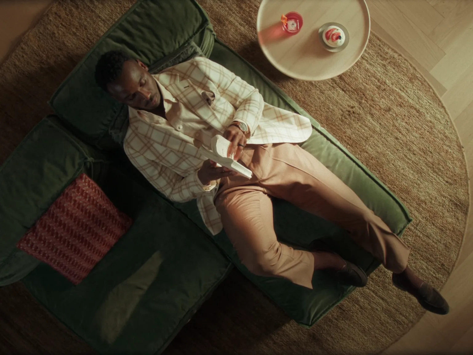 Homme allongé sur un canapé vert lisant un livre, avec une table ronde et des bougies à côté.
