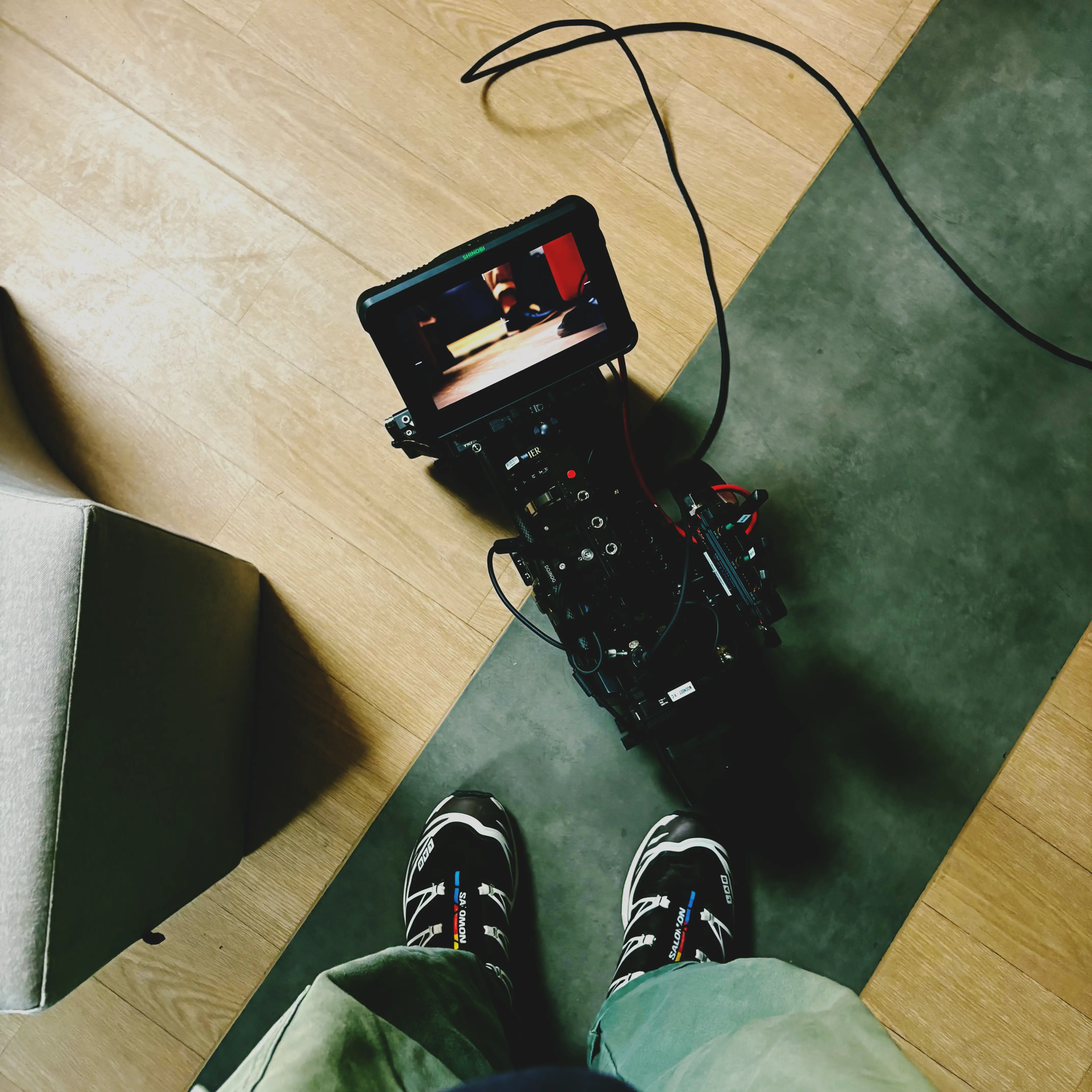 Vue plongeante d'une caméra vidéo professionnelle sur sol, avec deux pieds chaussés visibles.