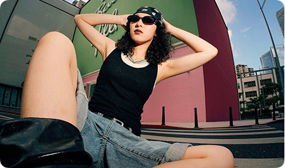 Person in sunglasses and bandana posing low-angle in urban setting with colorful walls and buildings.