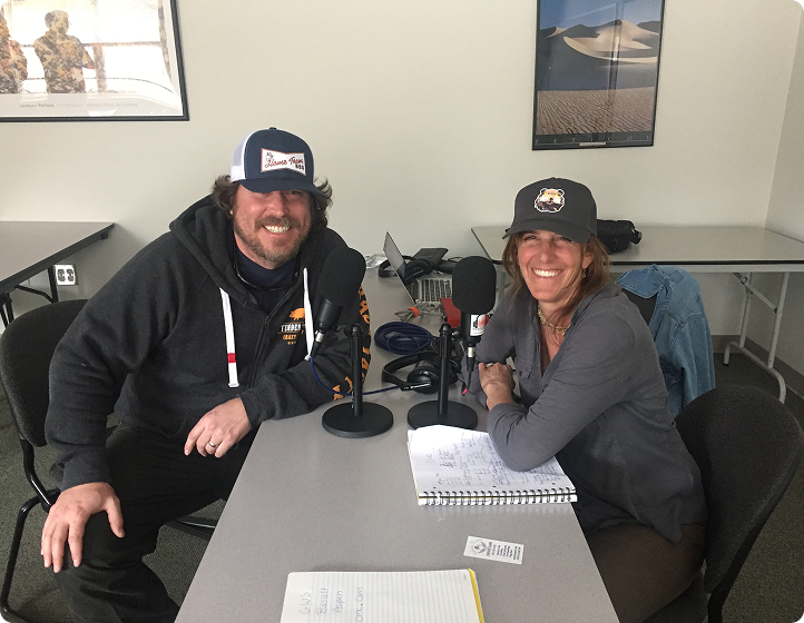 Two people wearing hats sitting at a table with microphones and notebooks, smiling at the camera.