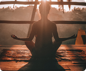 Silhouette of person meditating cross-legged on wooden floor with sunlight and nature backdrop.