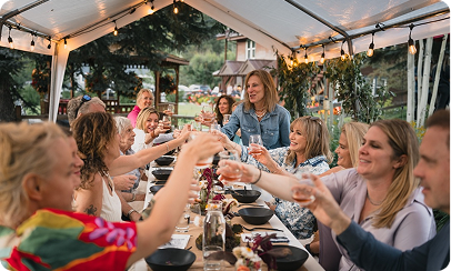 Group of people raising glasses for a toast at a festive outdoor dinner under a tent with string lights.