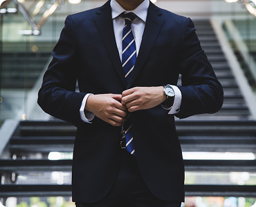 Man in dark suit with striped tie buttoning his jacket in an urban setting with stairs behind him.