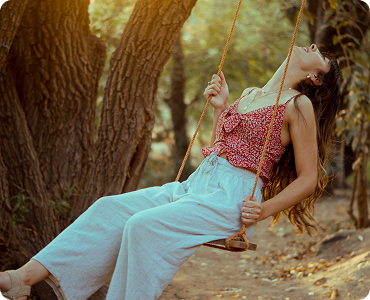 Young woman in red top swinging on a swing in a forest with trees and sunlight.