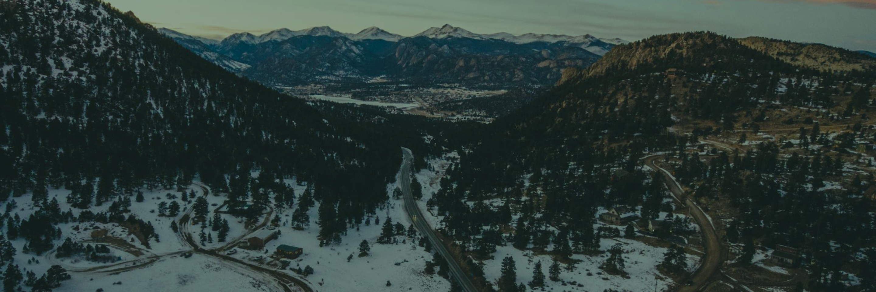 Snow-covered valley road winding through pine forests with mountains and a distant town in the background.