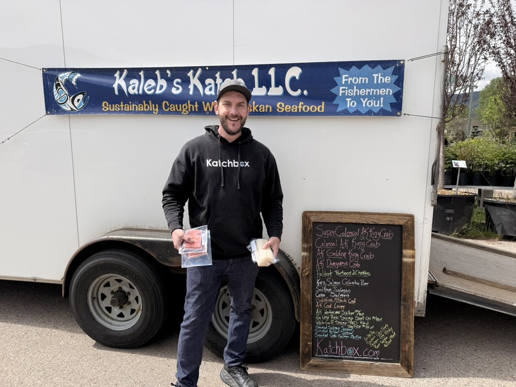 Man smiling holding seafood packages stands by a trailer with Kaleb's Katch LLC sign and seafood menu board.