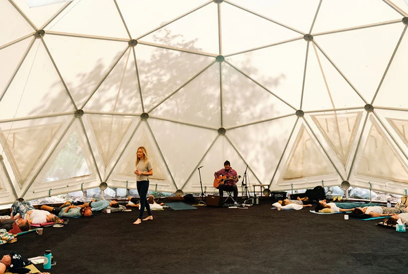 Woman stands barefoot inside dome while people lie on mats; man plays guitar nearby.