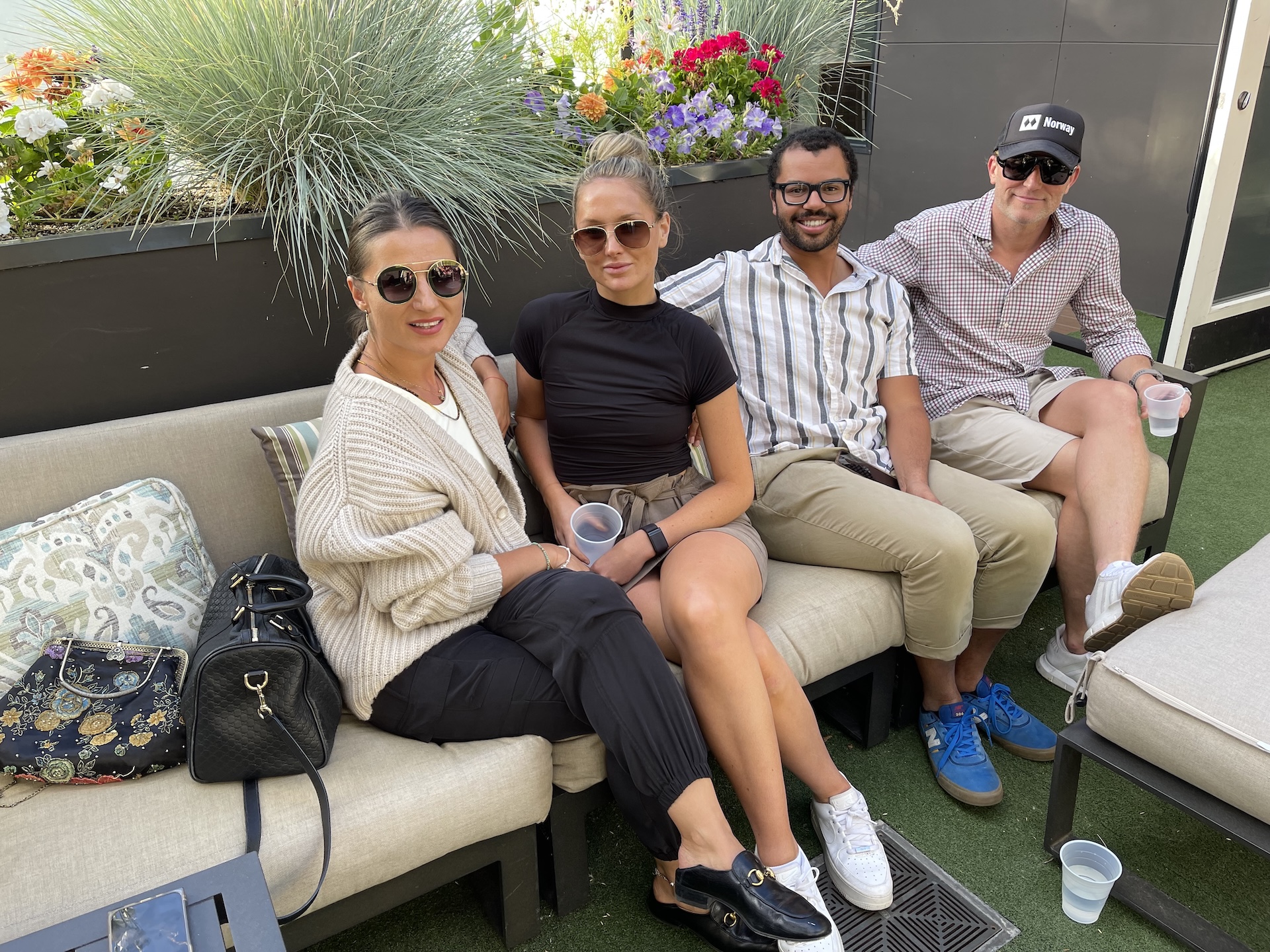 Two women and two men sitting on outdoor sofa with plants and flowers behind them.