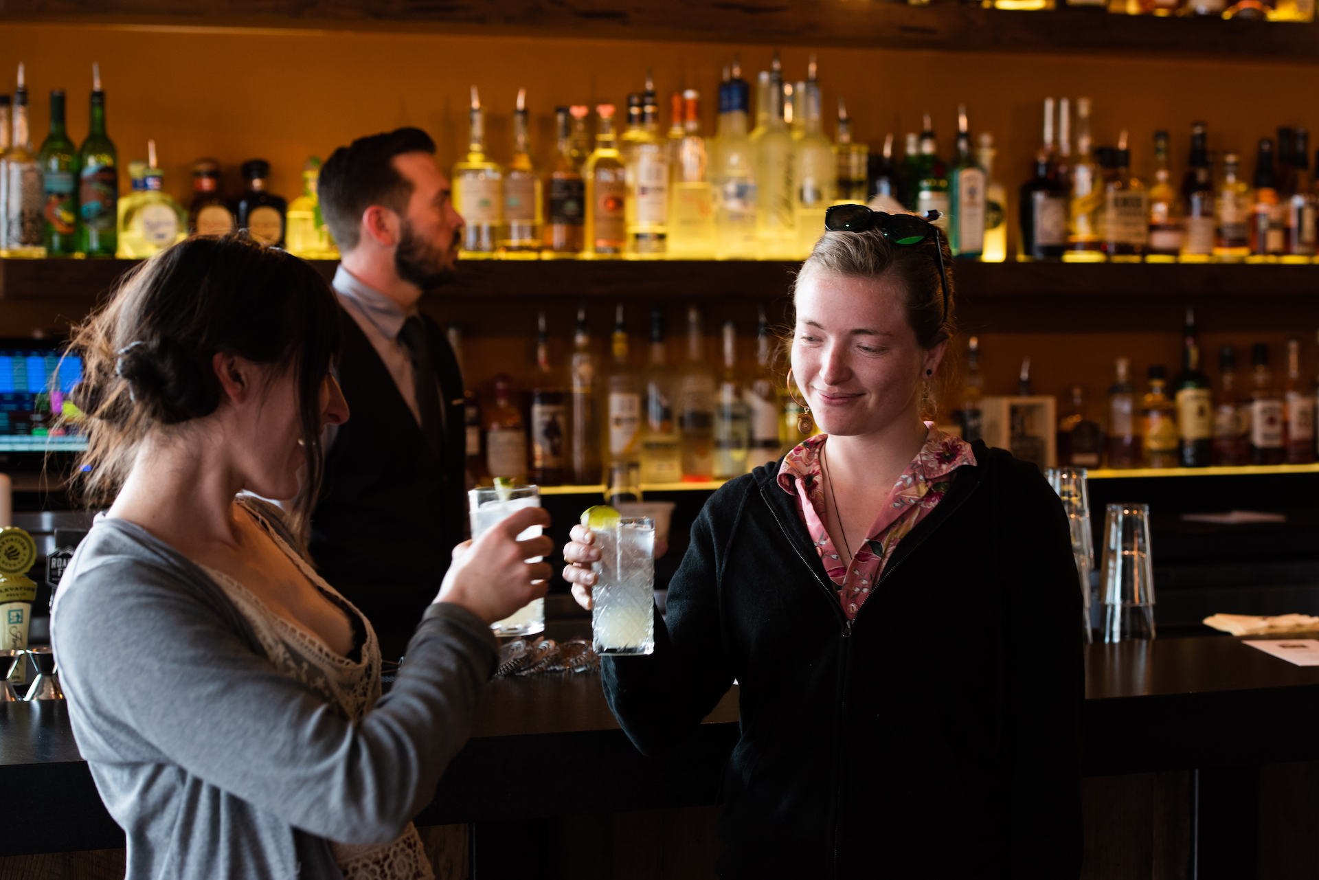 Two women clinking glasses with lime wedges in a bar with a bartender and liquor bottles in the background.