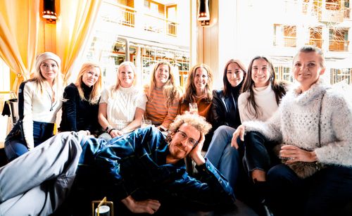 Group of eight women seated indoors with one man lying across their laps, all smiling at the camera.