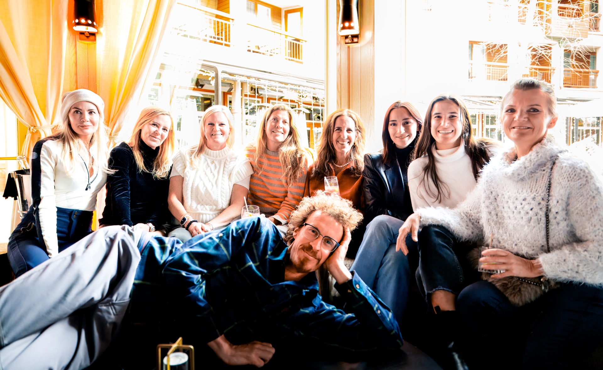 Group of eight women seated indoors with one man lying across their laps, all smiling at the camera.