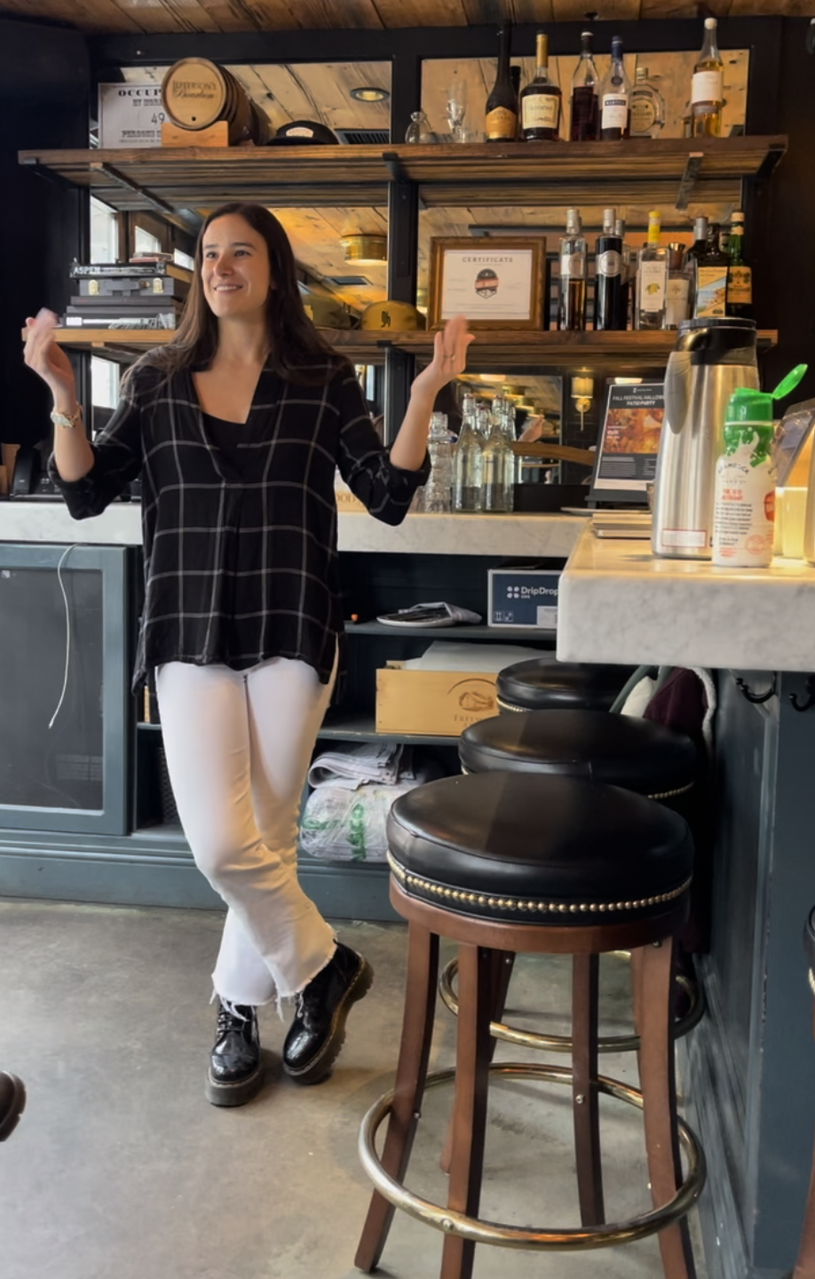 Woman with long dark hair wearing a black checkered shirt and white pants standing in a bar.