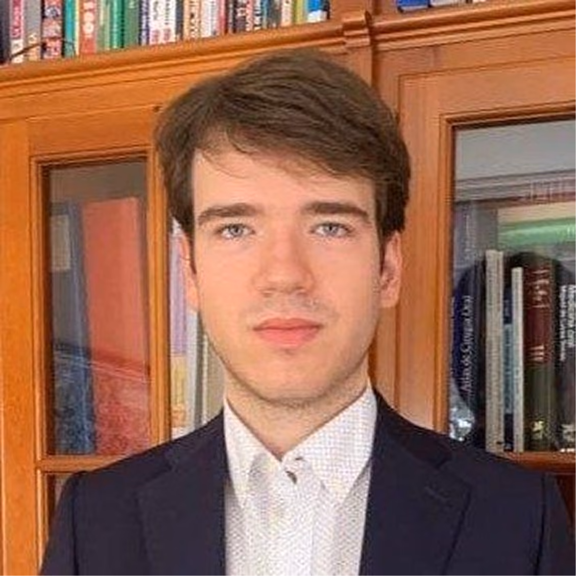 Young man with brown hair wearing a dark blazer and white shirt standing in front of a bookcase.
