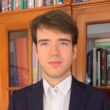 Young man in a dark blazer and white shirt standing in front of a wooden bookshelf with books.
